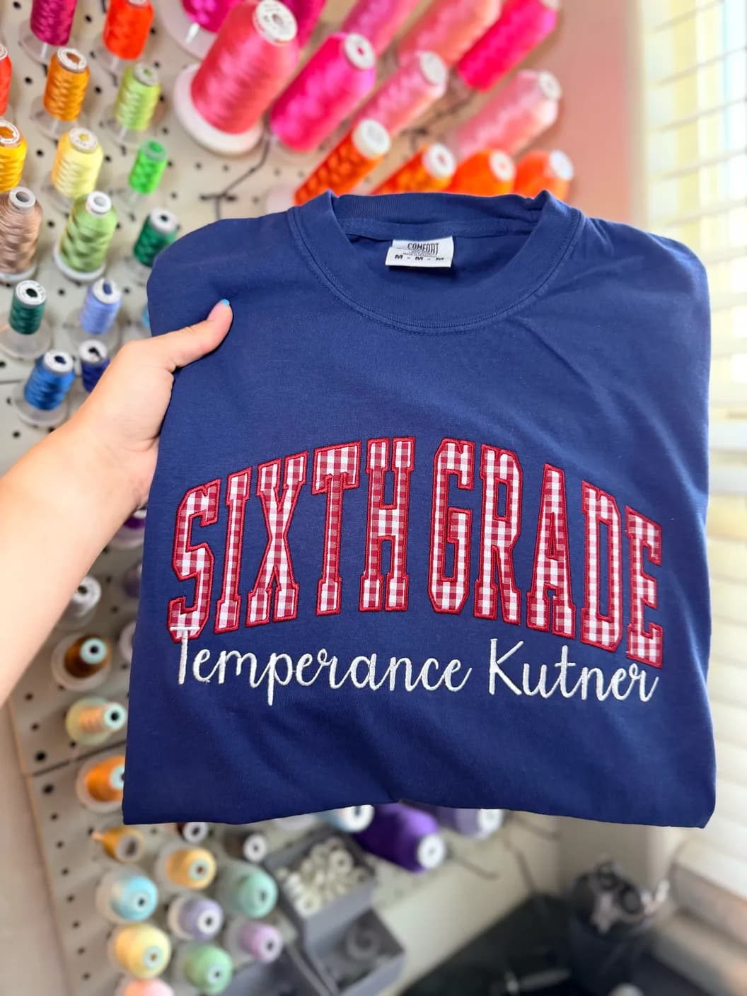 Navy shirt with red gingham Sixth Grade lettering and Temperance Kutner embroidery, held in front of colorful thread spools
