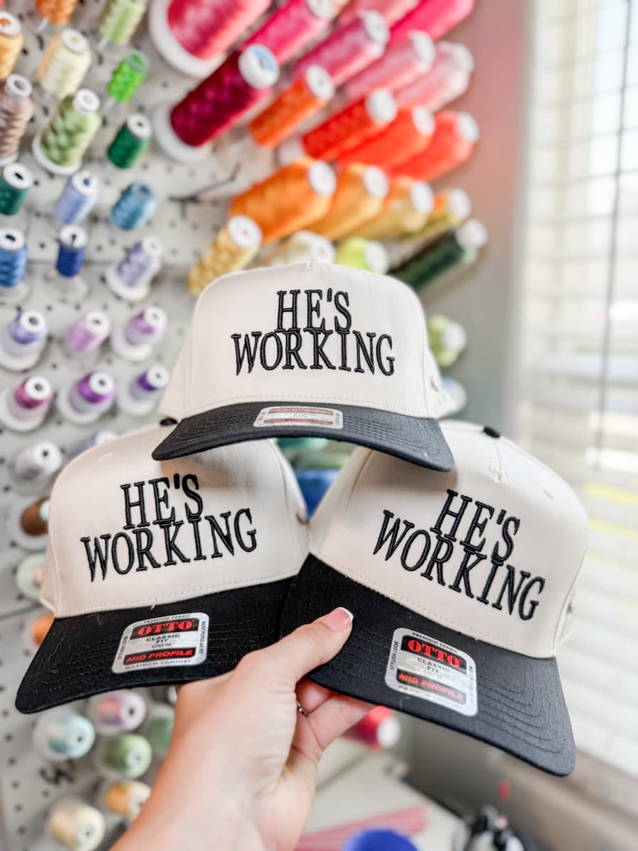 Custom embroidered trucker hats reading He's Working, held in front of a thread wall