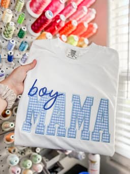 White tee with Boy Mama gingham applique lettering, held in front of a thread wall