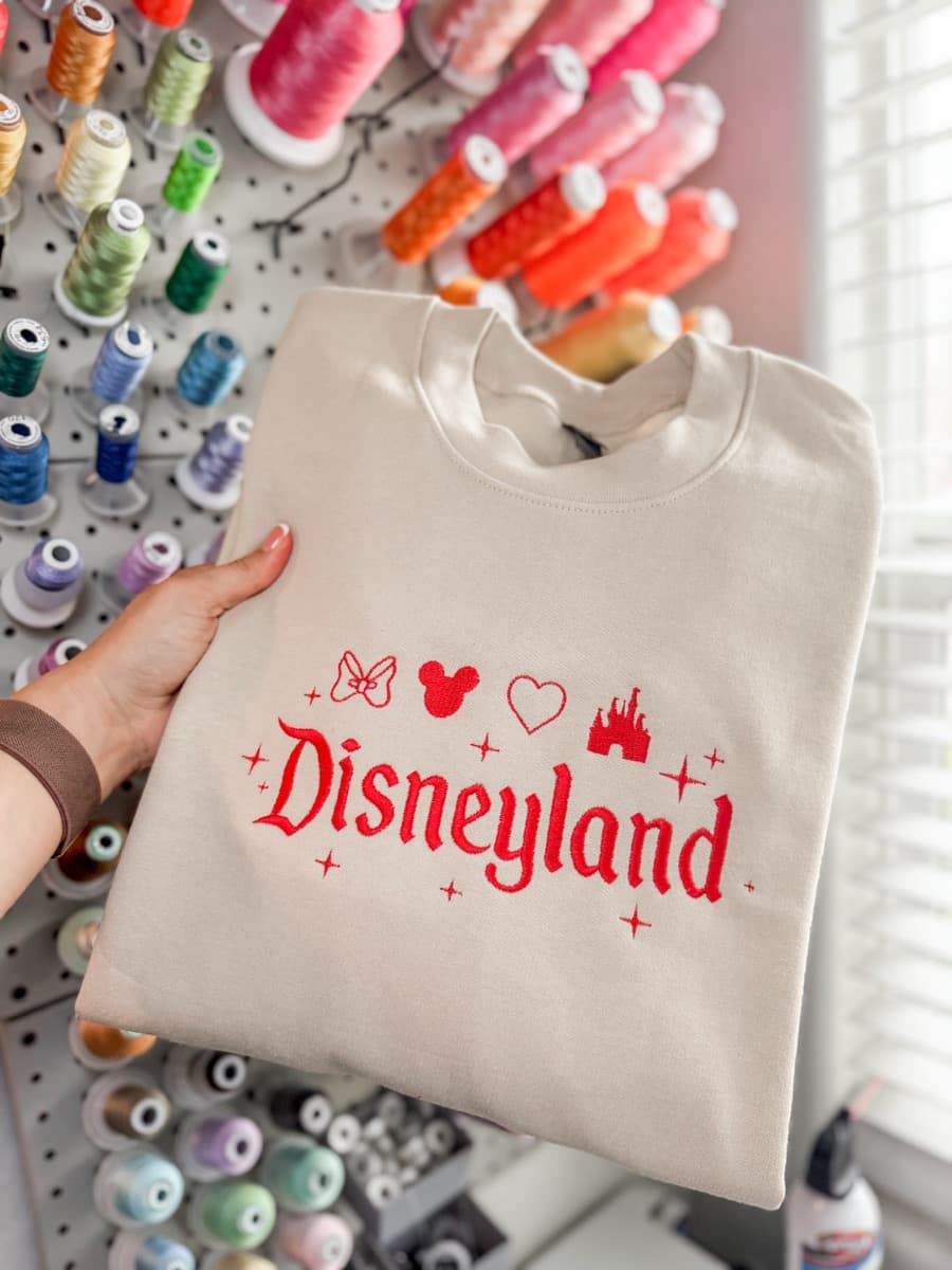 Cream sweatshirt with red Disneyland embroidery and Disney icons, held in front of a thread wall