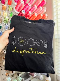 Black tee with dispatcher icons and script lettering in yellow, held in front of a thread wall