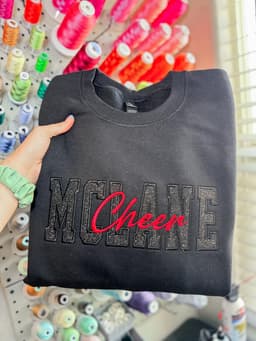Charcoal sweatshirt with McLane Cheer rhinestone lettering, held in front of a thread wall