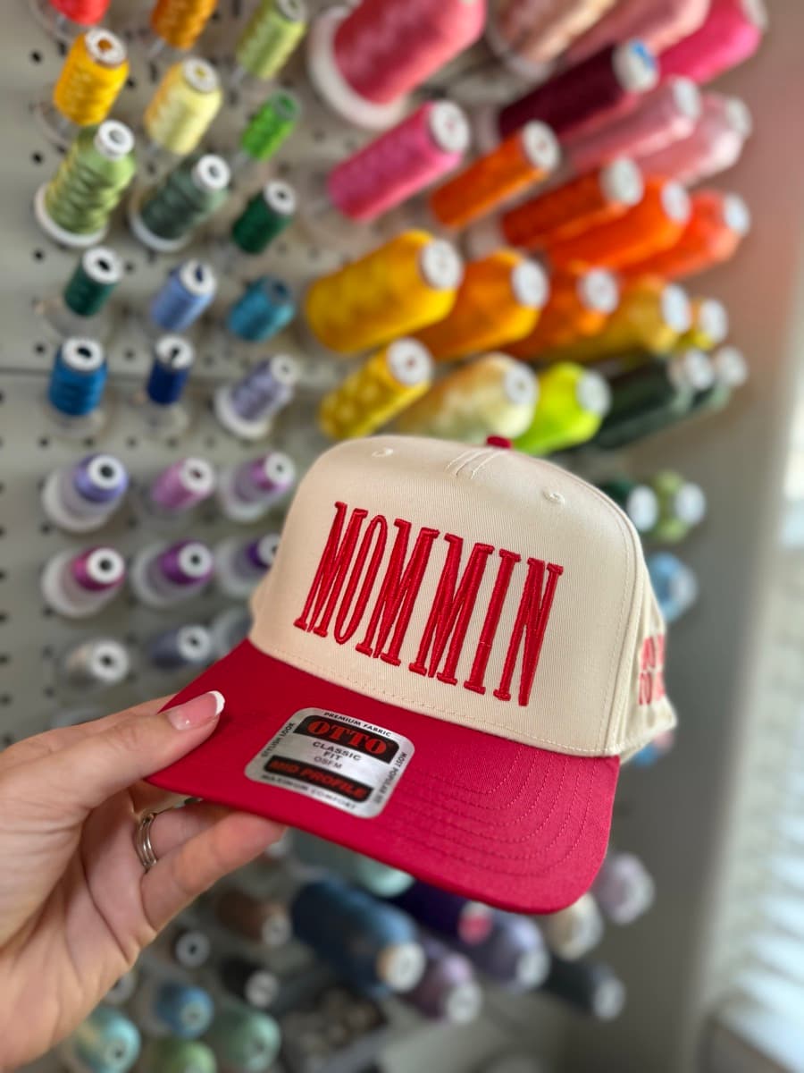 Cream and red trucker hat with MOMMIN embroidery, held in front of a thread wall