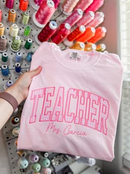 Pink tee with Teacher varsity lettering personalized with a name, held in front of a thread wall