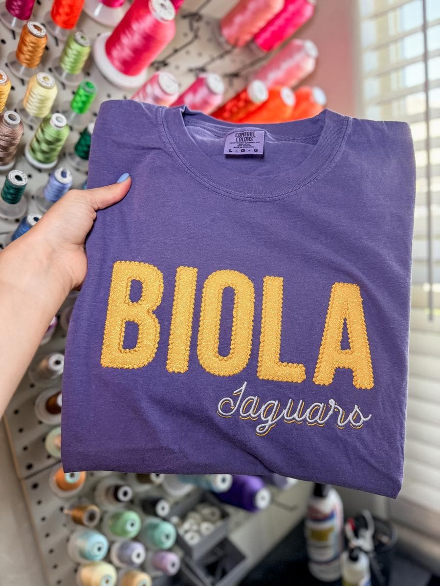 Purple tee with Biola Jaguars chenille lettering, held in front of a thread wall