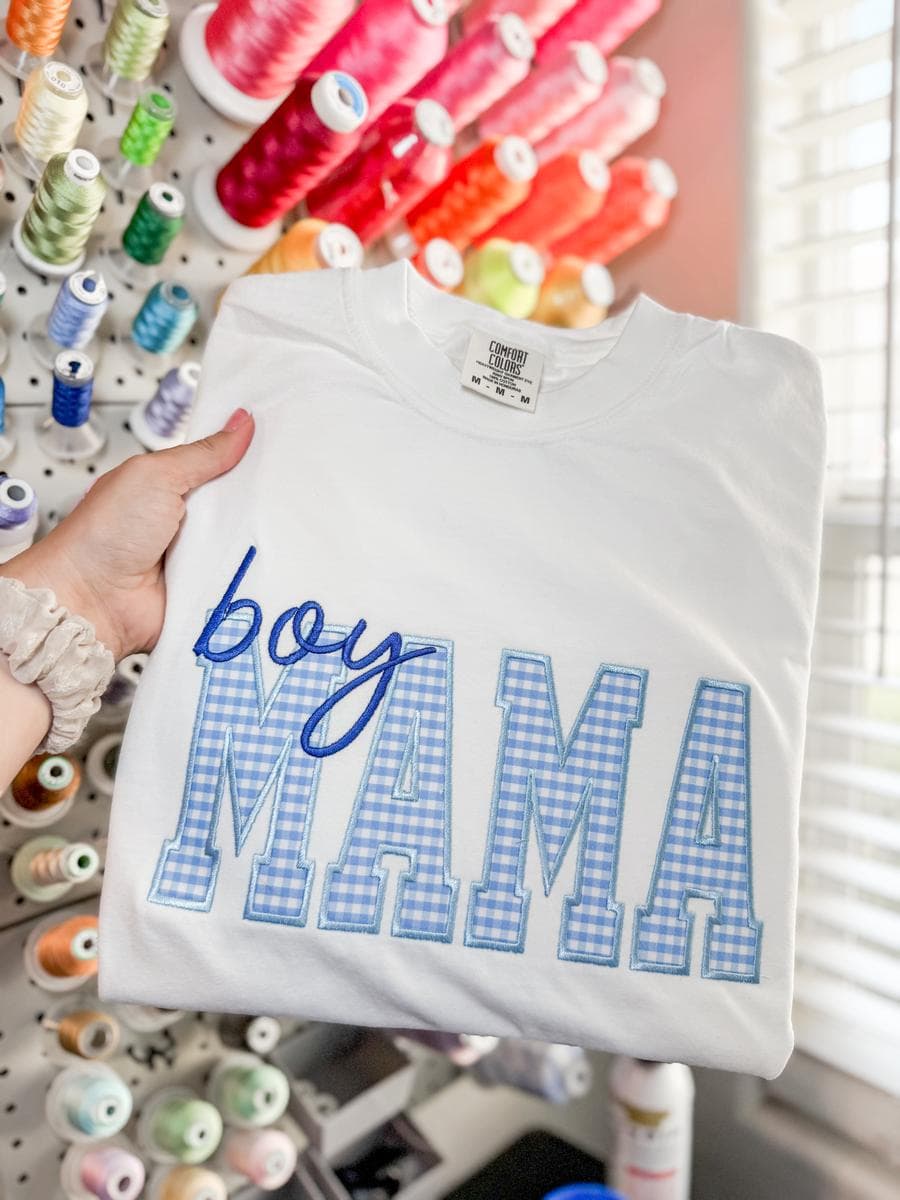 White tee with Boy Mama gingham applique lettering, held in front of a thread wall