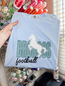Light blue tee with Broncos football lettering and horse embroidery, held in front of a thread wall