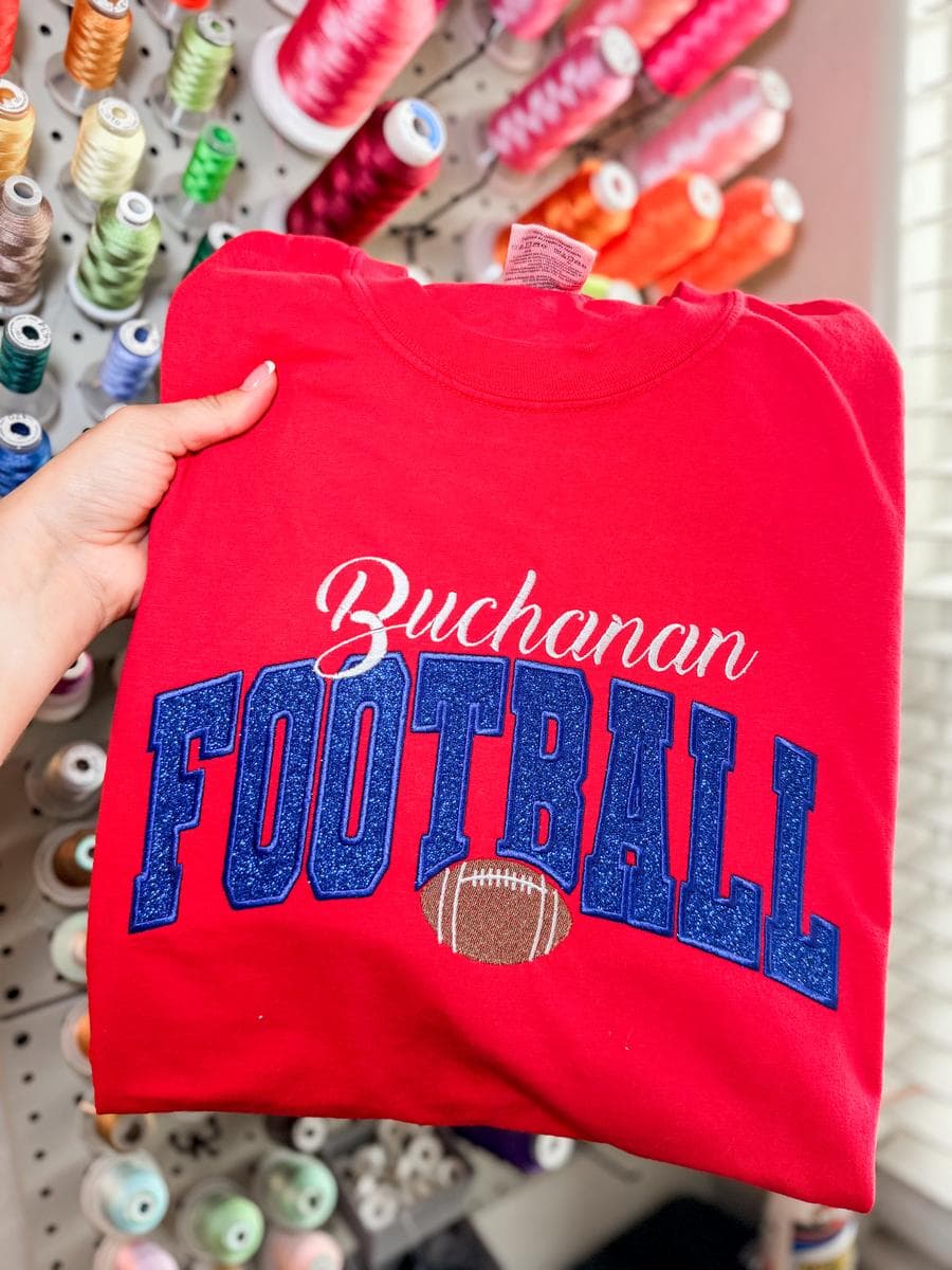 Red tee with Buchanan Football glitter lettering, held in front of a thread wall
