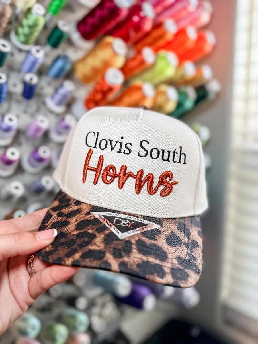 Cream trucker hat with Clovis South Horns embroidery and leopard print brim, held in front of a thread wall