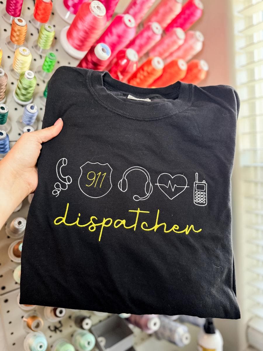 Black tee with dispatcher icons and script lettering in yellow, held in front of a thread wall