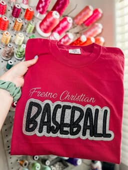 Red tee with Fresno Christian Baseball applique lettering, held in front of a thread wall