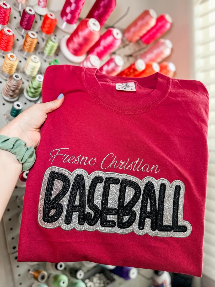 Red tee with Fresno Christian Baseball applique lettering, held in front of a thread wall