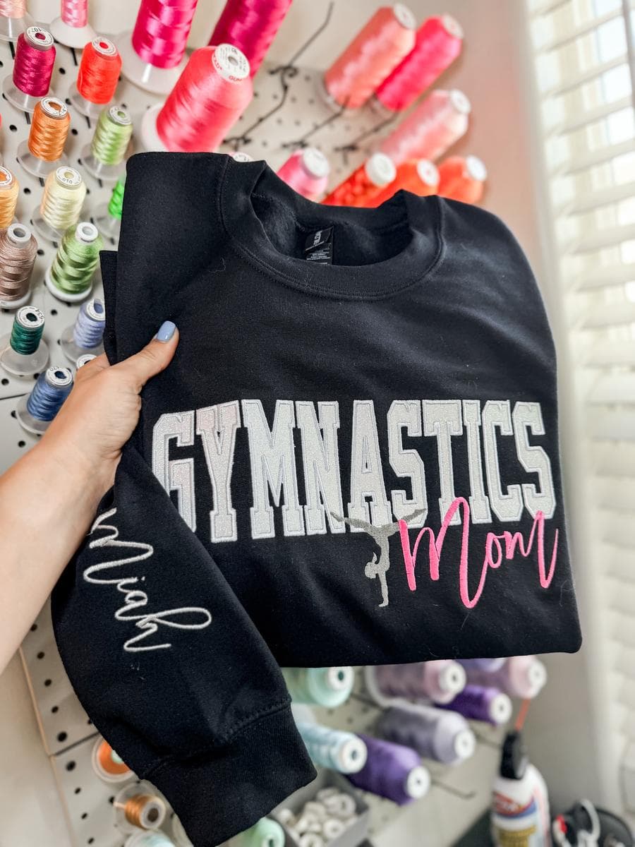 Black sweatshirt with Gymnastics Mom glitter lettering, held in front of a thread wall