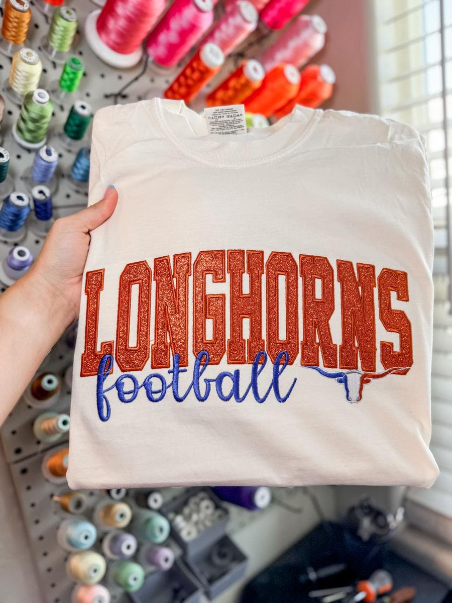 White tee with Longhorns football glitter and script lettering, held in front of a thread wall