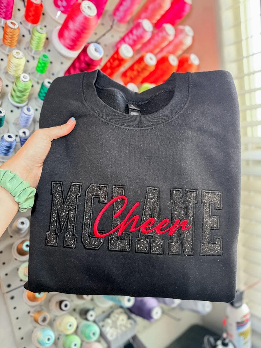 Charcoal sweatshirt with McLane Cheer rhinestone lettering, held in front of a thread wall