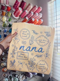 Custom DTF print on a pale yellow tee with nana lettering and floral illustrations, held in front of a thread wall