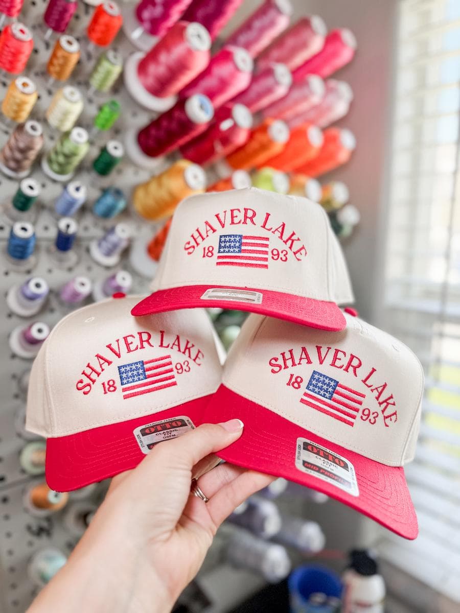Three red and white trucker hats with Shaver Lake flag embroidery, held in front of a thread wall